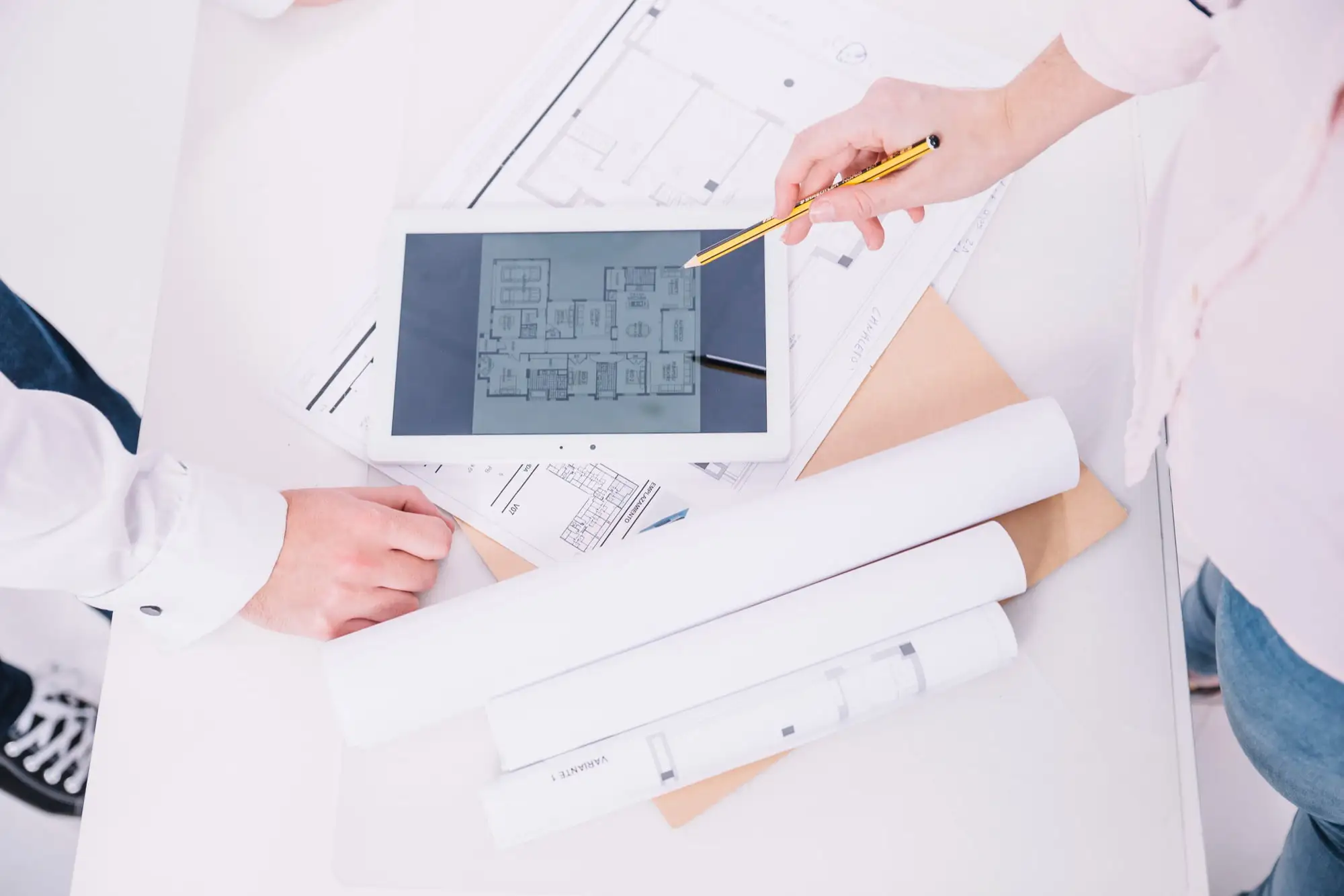 Two professionals reviewing architectural plans on a tablet and paper documents, with rolled blueprints on the desk