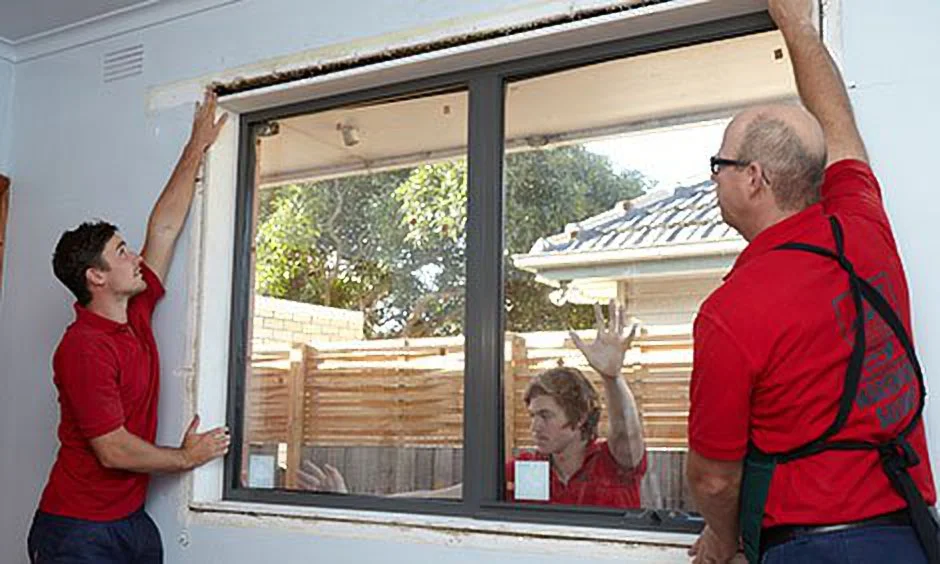 Two workers in red shirts install a window in a home